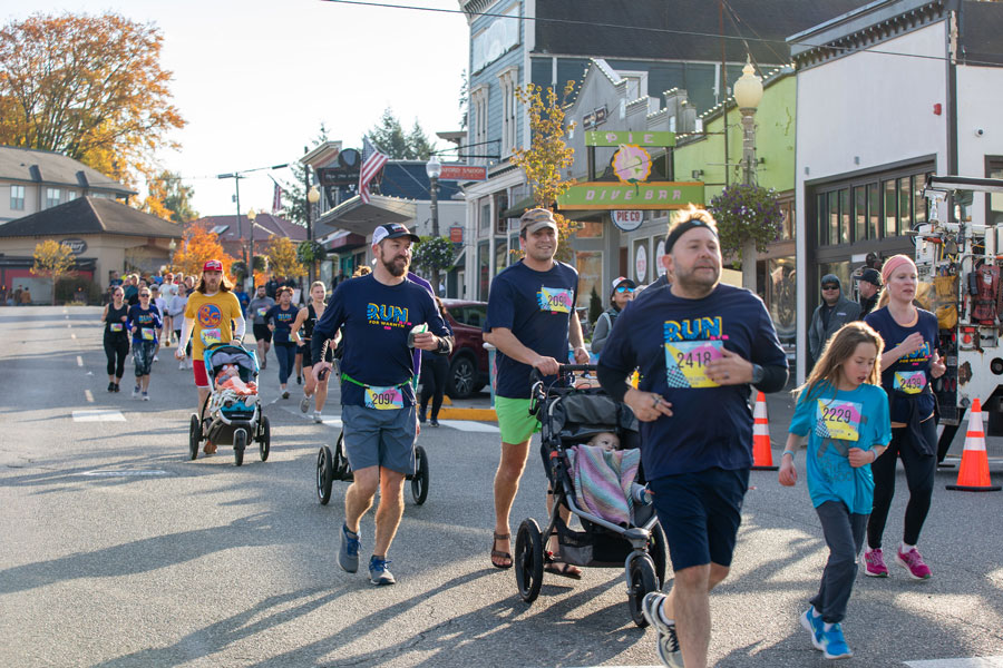 A trio of men in dark colors run in the Run for Warmth. One is smiling and pushing a stroller.