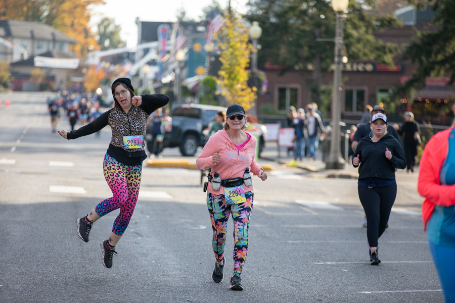 Two women in brightly colored leggings celebrate participation in the Run for Warmth. One is leaping into the air while the other smiles and runs toward the camera.