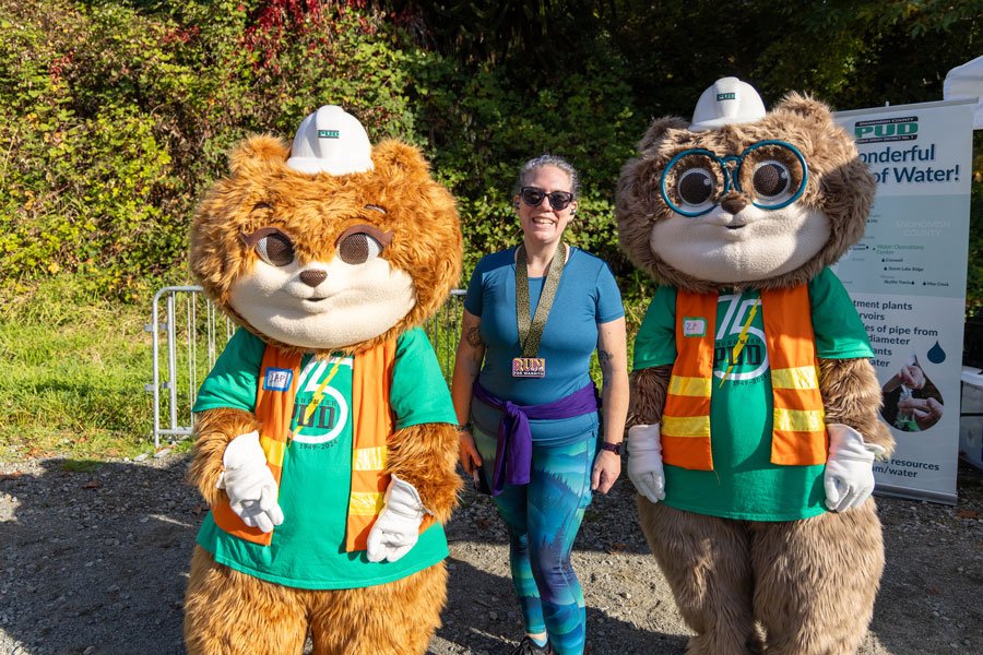 Zip and Zap pose with a smiling racer after she finished the Run for Warmth. She wears a medal and Zip and Zap are in bright vests and anniversary shirts.